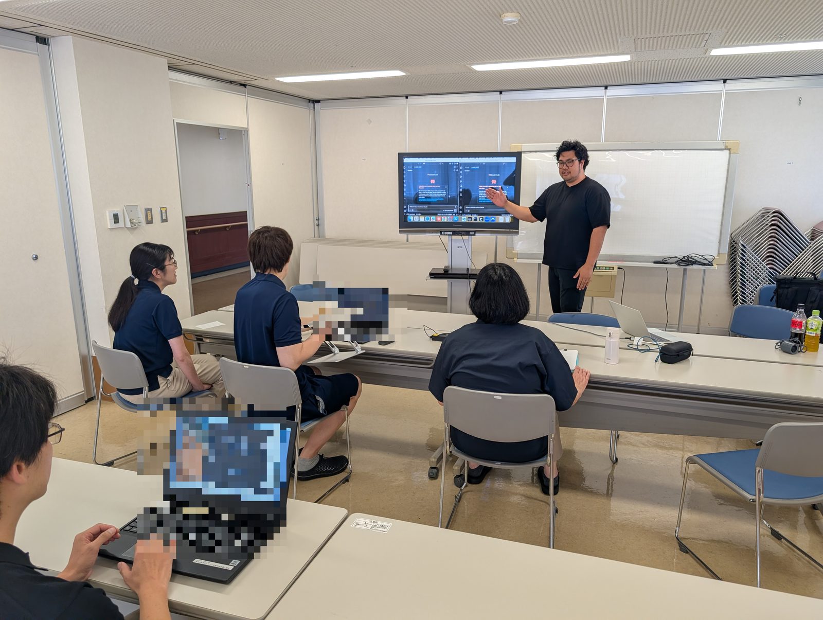 福祉施設の一室で、制服姿の職員数名がノートパソコンを開き、合同会社サンの代表による研修を受けている。前方の大画面にはAIツールの画面が映っている。